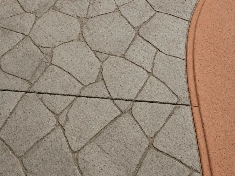 Close-up of stamped concrete in a gray random flagstone pattern meeting a smooth terracotta border band, with a saw-cut joint across the slab